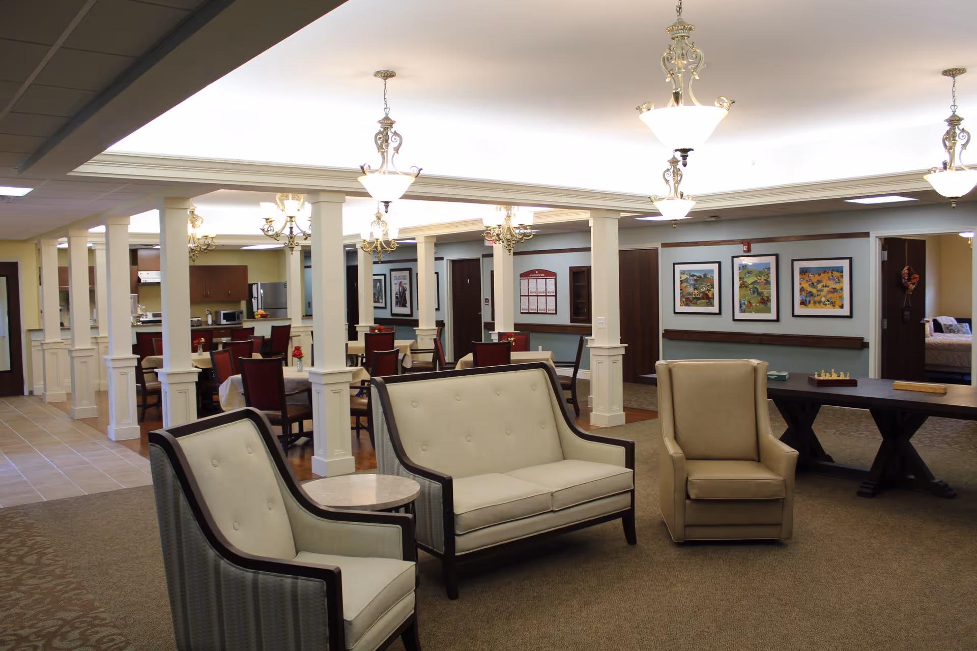 A spacious and well-lit common area in a senior living facility featuring comfortable seating including two armchairs and a loveseat arranged around a small round table. In the background, there is a dining area with multiple tables and chairs, decorative columns, framed artwork on the walls, and a kitchen area with appliances. The ceiling has elegant hanging light fixtures.