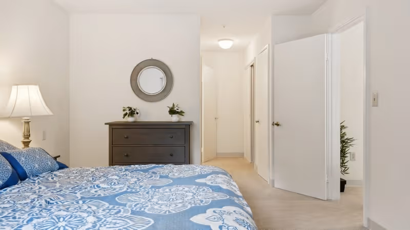 A bedroom with a bed covered in a blue and white patterned bedspread, a bedside lamp on a nightstand, a dark wooden dresser with two small plants on top, and a round mirror hanging above the dresser. The room has white walls and carpeted floor, with an open doorway leading to a hallway and another room.