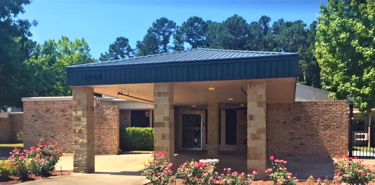 Front entrance of a single-story brick building with a covered drop-off area supported by stone pillars, surrounded by greenery and flowering bushes under a clear blue sky.