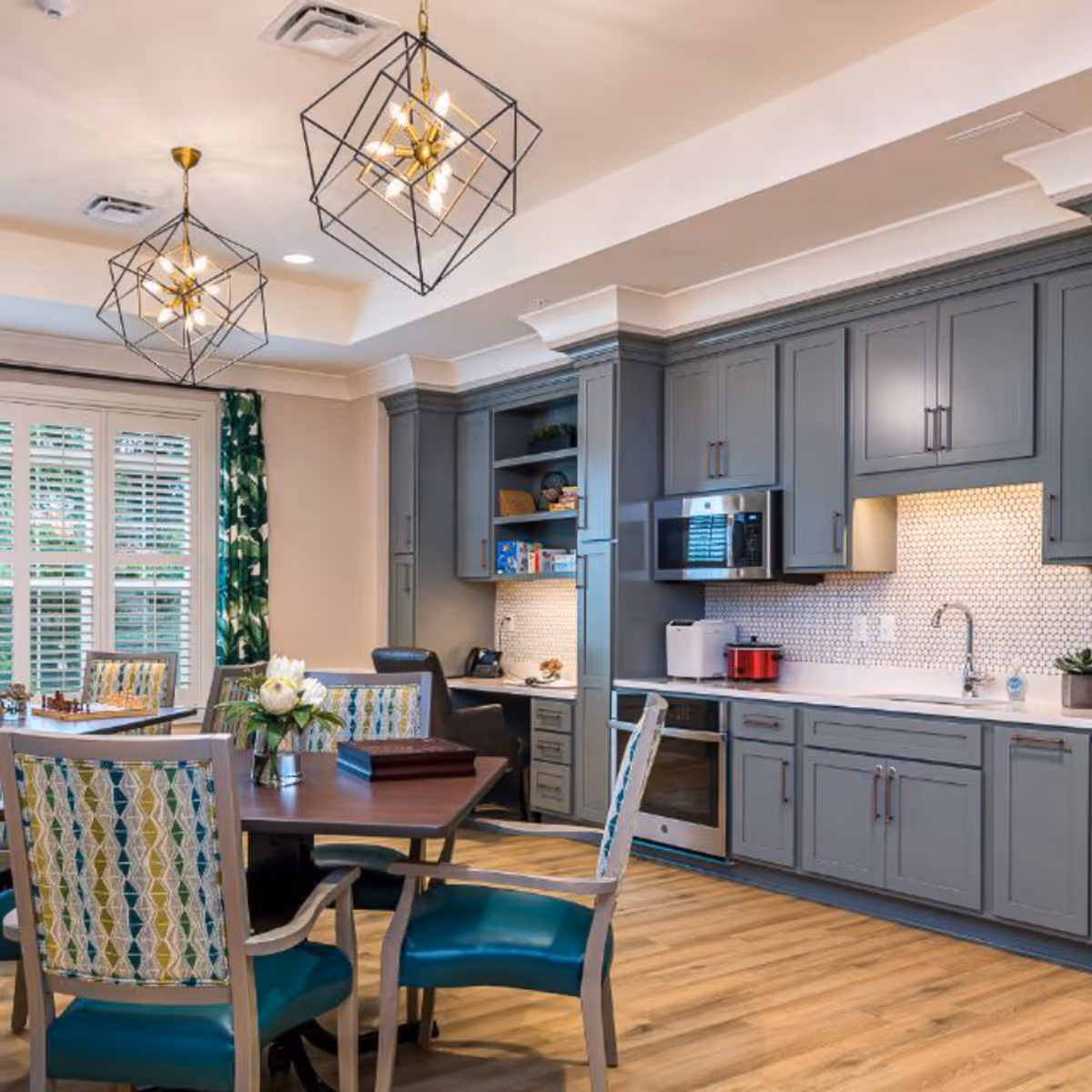 A modern kitchen and dining area with gray cabinets, a white countertop, a built-in microwave, and a stove. The dining area has a wooden table with four chairs featuring patterned upholstery and teal seats. Two geometric pendant lights hang from the ceiling, and there are large windows with white shutters and green patterned curtains.
