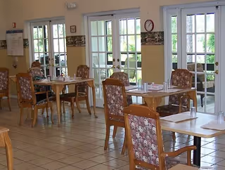 Dining area with several wooden tables and floral upholstered chairs. Large glass doors and windows let in natural light, and a person is seated at one of the tables. The room has tiled floors and light-colored walls with a decorative border.