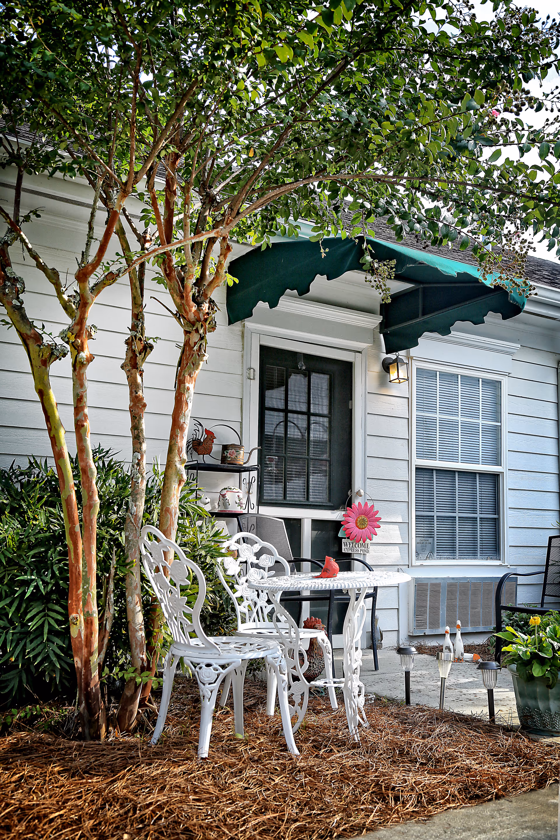 Outdoor seating area with white metal table and chairs under a tree next to a building entrance with a green awning. There are decorative items including a pink flower welcome sign, a small red bird figurine on the table, and garden ornaments near the window and plants.