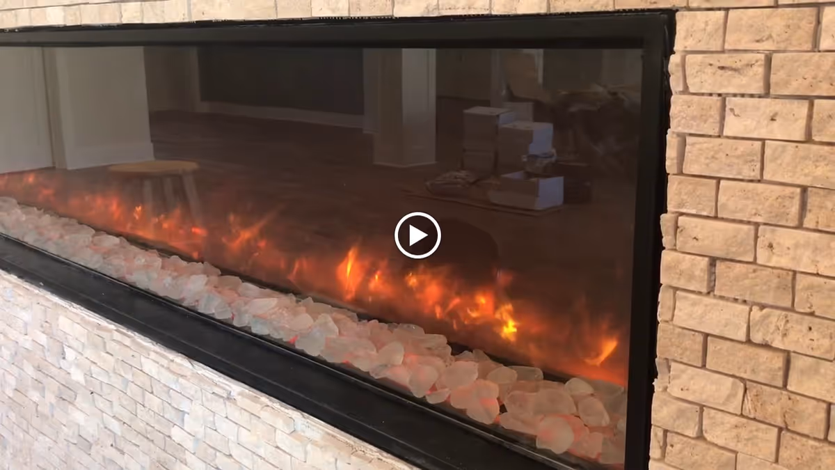 A modern electric fireplace with glowing embers and white decorative stones inside a stone and brick surround. The reflection in the glass shows a room with wooden flooring, a stool, and some stacked boxes.