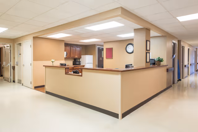 Reception desk area inside a nursing and rehabilitation center with beige walls, a clock on the wall, wooden cabinets, a white refrigerator, and a hallway with doors leading to other rooms.