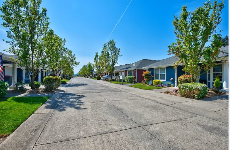A wide concrete street lined with single-story residential buildings on both sides, each with small front yards featuring green grass, bushes, and trees under a clear blue sky.