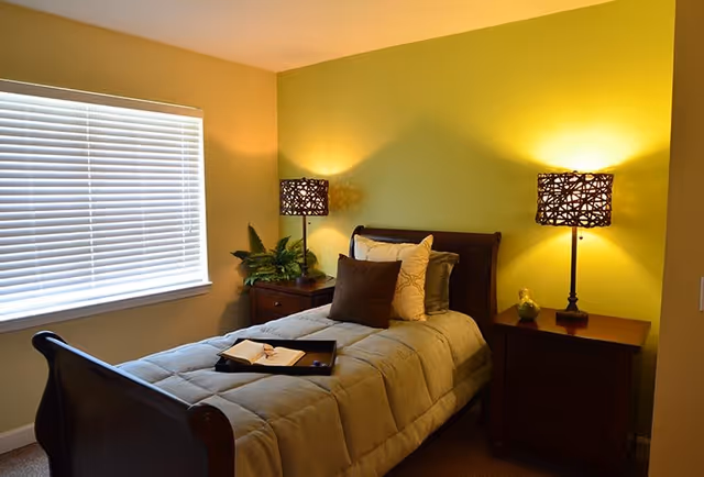 A cozy bedroom with a single bed featuring a gray comforter and multiple pillows. There are two wooden nightstands on either side of the bed, each with a decorative lamp emitting warm light. A window with closed blinds is on the left wall, and a small plant is placed on the left nightstand.