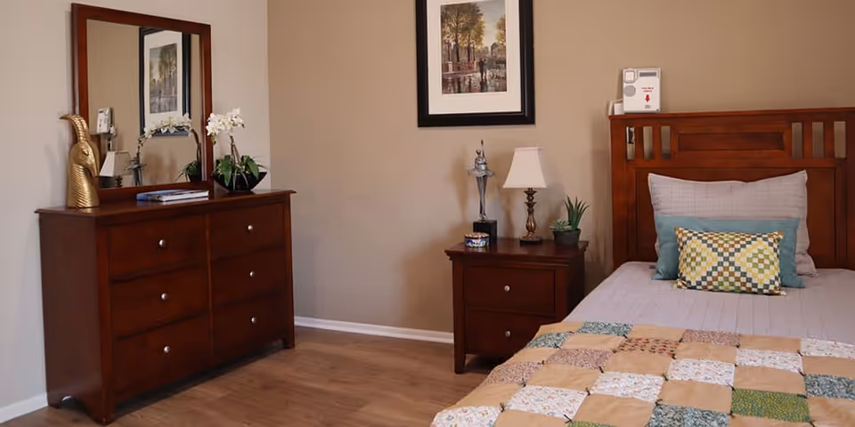 A cozy bedroom with a wooden bed featuring a patchwork quilt and decorative pillows. Next to the bed is a wooden nightstand with a lamp, a small plant, and a decorative item. On the opposite wall is a wooden dresser with six drawers, a large mirror, and decorative items including a golden bird sculpture and a potted orchid. The walls are beige and there is a framed picture hanging above the nightstand.