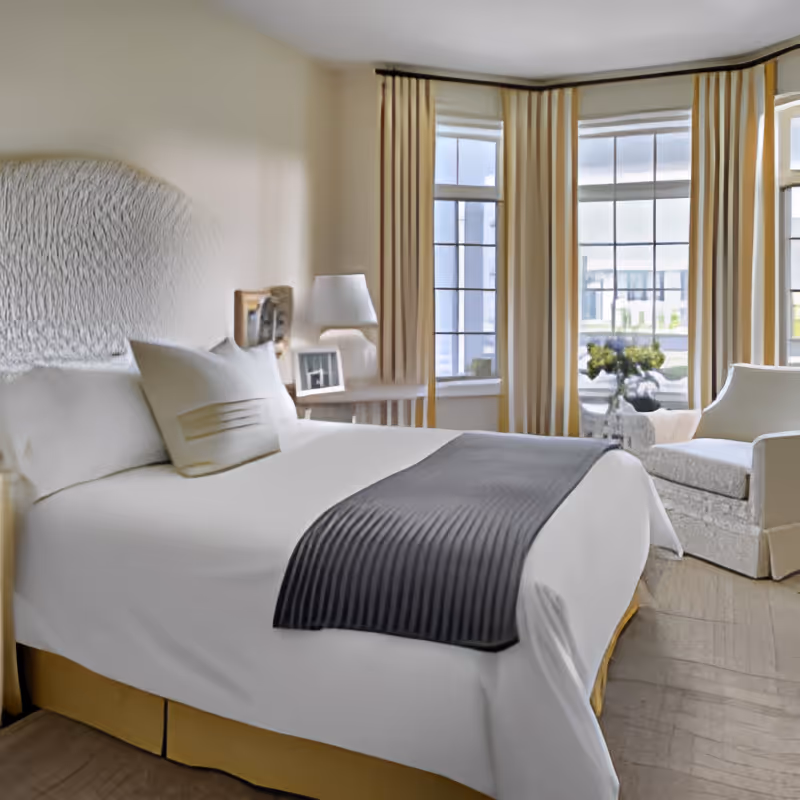 A bright and cozy bedroom with a large bed featuring white linens and a dark gray throw blanket. The room has a textured headboard, a bedside table with a lamp and framed photos, large windows with beige curtains letting in natural light, and a comfortable armchair next to a small table with a vase of flowers.
