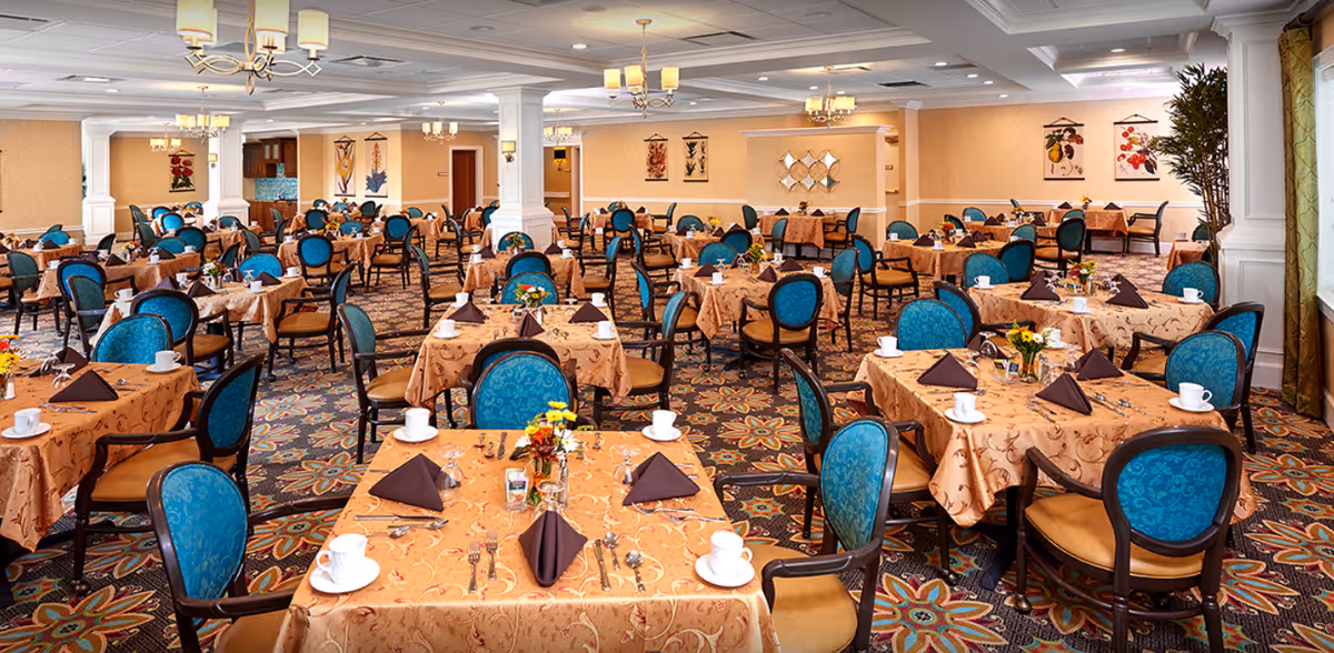 Spacious, elegantly decorated dining room with many tables set with tablecloths, teal upholstered chairs, and place settings.