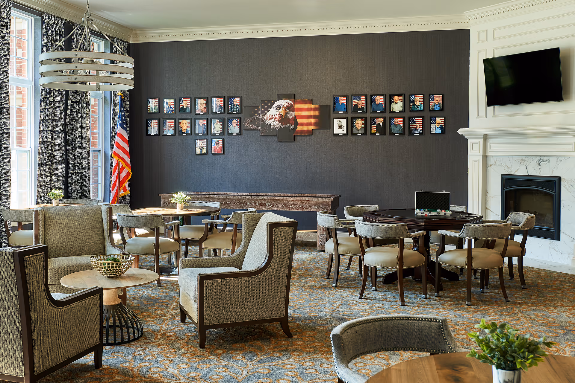 A comfortable and well-lit common area with multiple seating arrangements including armchairs and round tables with chairs. The room features a large wall decorated with framed photos and an American flag, a fireplace with a mounted TV above it, and large windows with curtains allowing natural light to enter.