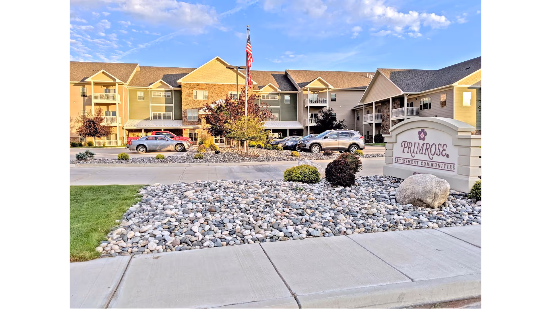 Exterior view of Primrose Retirement Community of Gillette showing a multi-story building with balconies, parked cars, an American flag on a flagpole, and a landscaped area with rocks and bushes in front of a sign displaying the community's name.