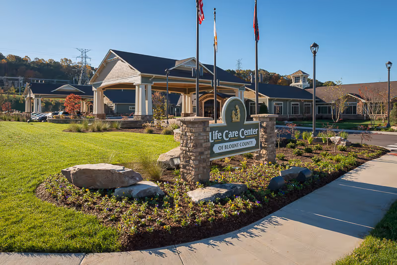 Exterior view of Life Care Center of Blount County showing the main entrance with a covered drop-off area, a stone sign with the facility's name, landscaped garden beds with rocks and flowers, a well-maintained lawn, and several flagpoles with flags. The building is single-story with a dark roof and beige siding, set against a backdrop of trees and a clear blue sky.