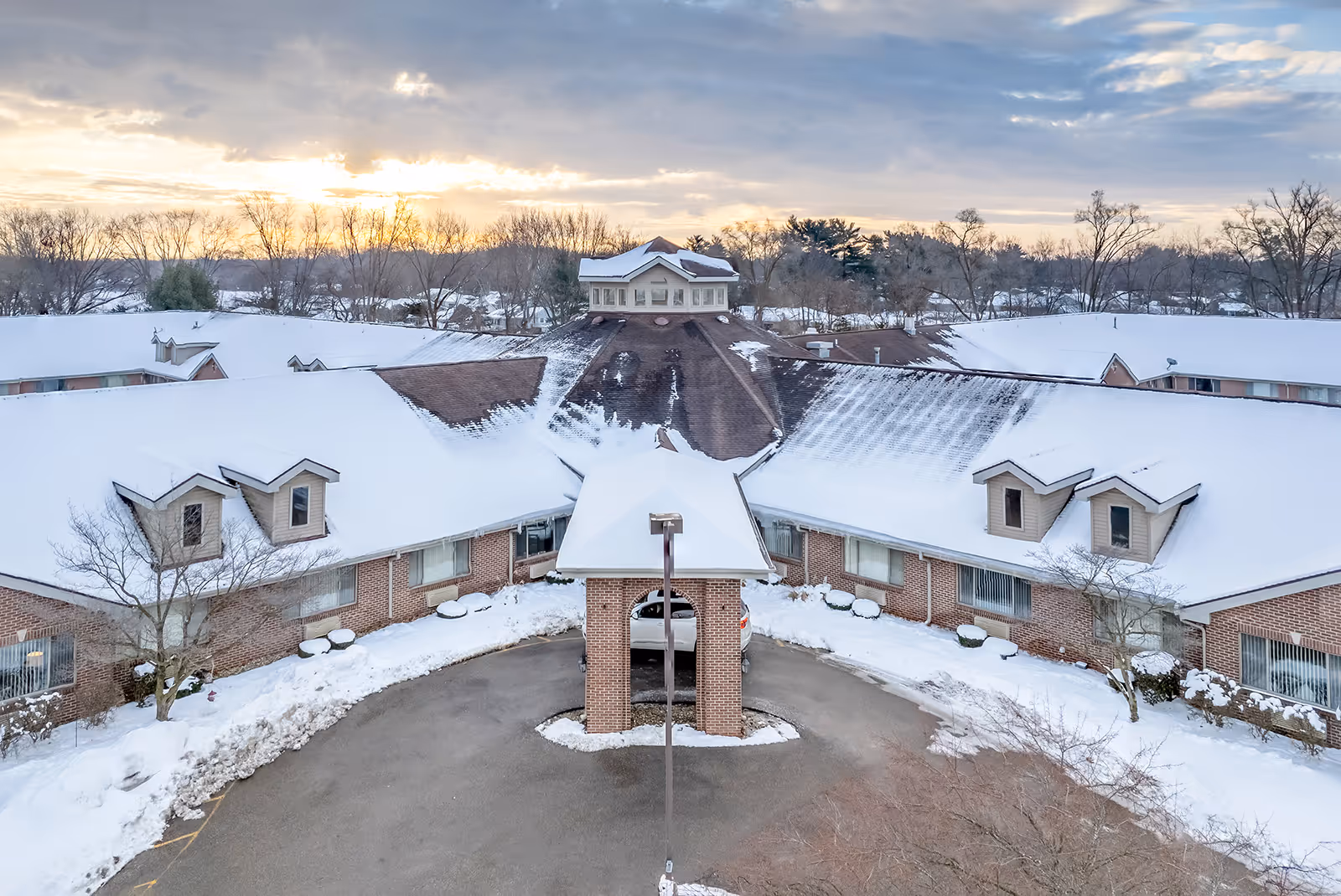 Aerial view of Brentwood at Elkhart Assisted Living facility covered in snow during sunset, showing a circular driveway with a covered entrance and surrounding brick buildings with snow-covered roofs and leafless trees in the background.