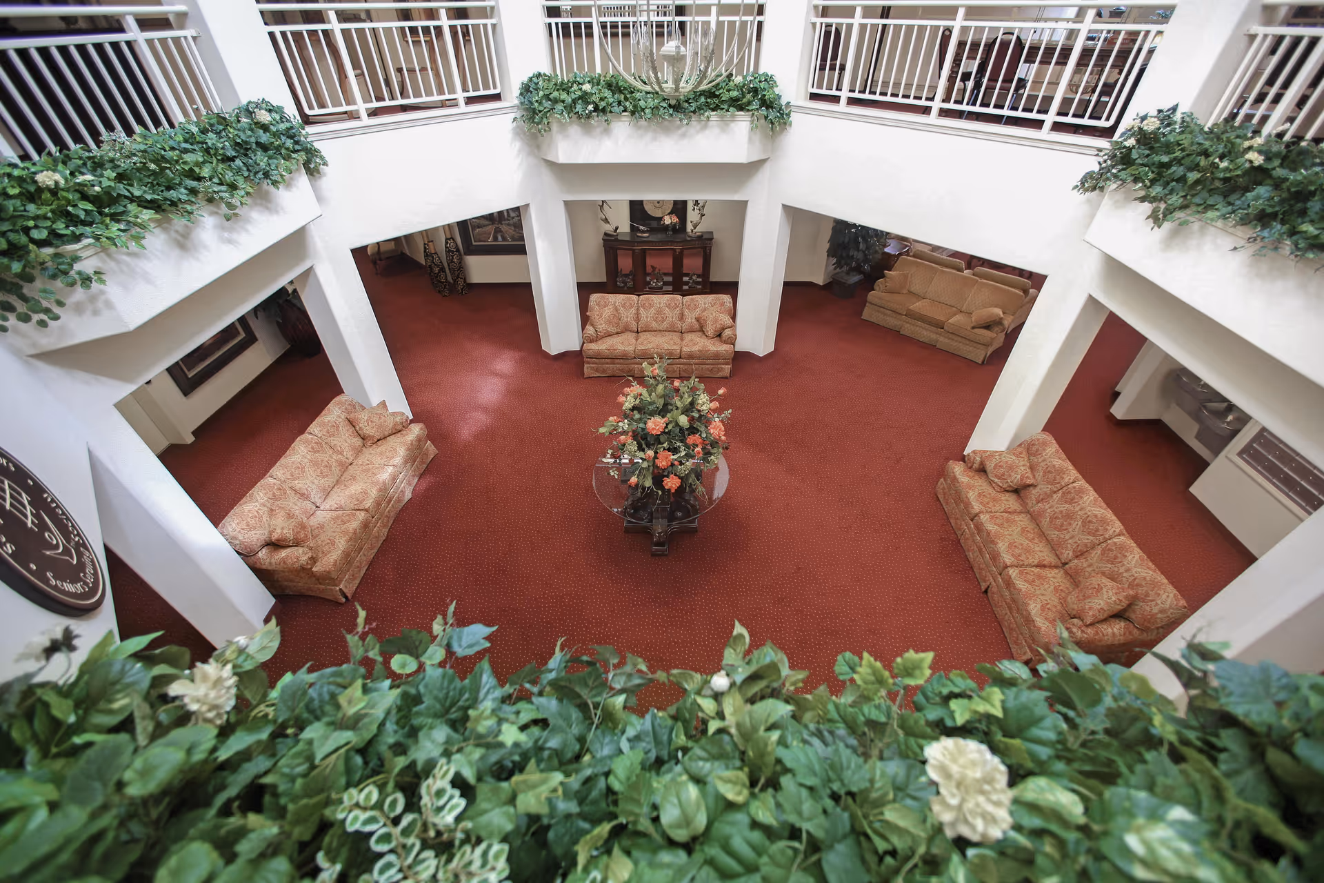 View from an upper level looking down into a spacious common area with red carpet, four patterned sofas arranged around a central glass table with a floral arrangement, white walls, and green plants along the balcony railing.