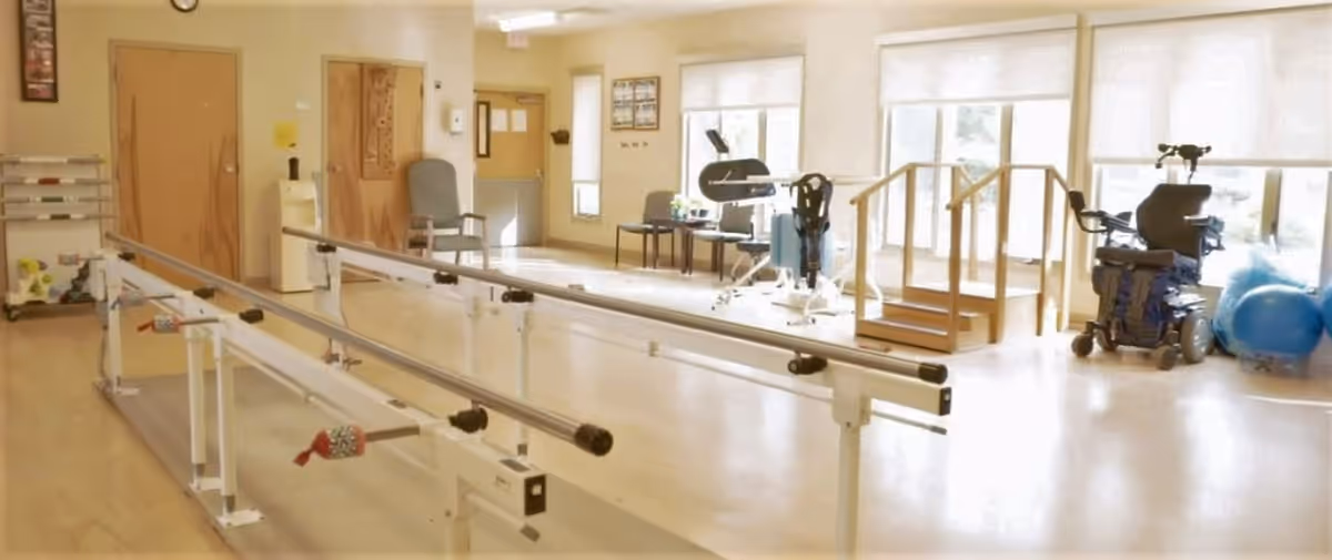 A bright rehabilitation room with parallel bars for walking exercises, chairs, exercise equipment, a small wooden staircase with handrails, and a power wheelchair near large windows letting in natural light.