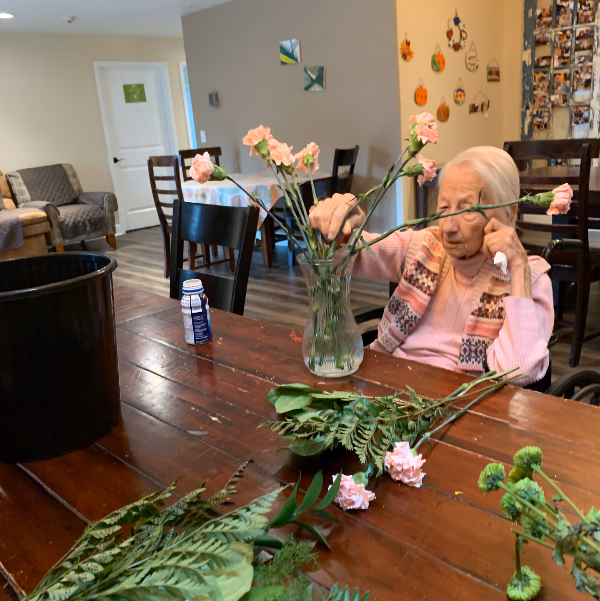 An elderly woman sitting at a wooden table arranging pink flowers in a glass vase inside a cozy room with chairs, a couch, and wall decorations.