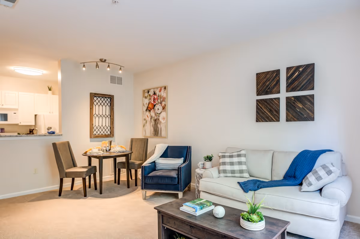 A cozy living room and dining area in a senior living facility. The living room features a light-colored sofa with patterned pillows and a blue throw blanket, a dark blue armchair with a striped pillow, and a wooden coffee table with books and a small plant. The dining area has a small round table set for two with two upholstered chairs. The kitchen is visible in the background with white cabinets and appliances. The walls are decorated with floral and wooden artwork.