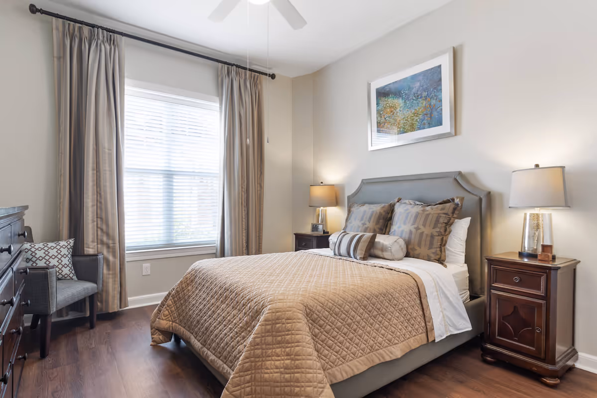 A neatly made bed with a beige quilted bedspread and multiple decorative pillows in a bedroom. The room features a window with beige curtains, two wooden nightstands with lamps on either side of the bed, a framed artwork on the wall above the headboard, a wooden dresser, and a gray upholstered chair with a patterned pillow. The floor is wooden and the walls are painted light gray.