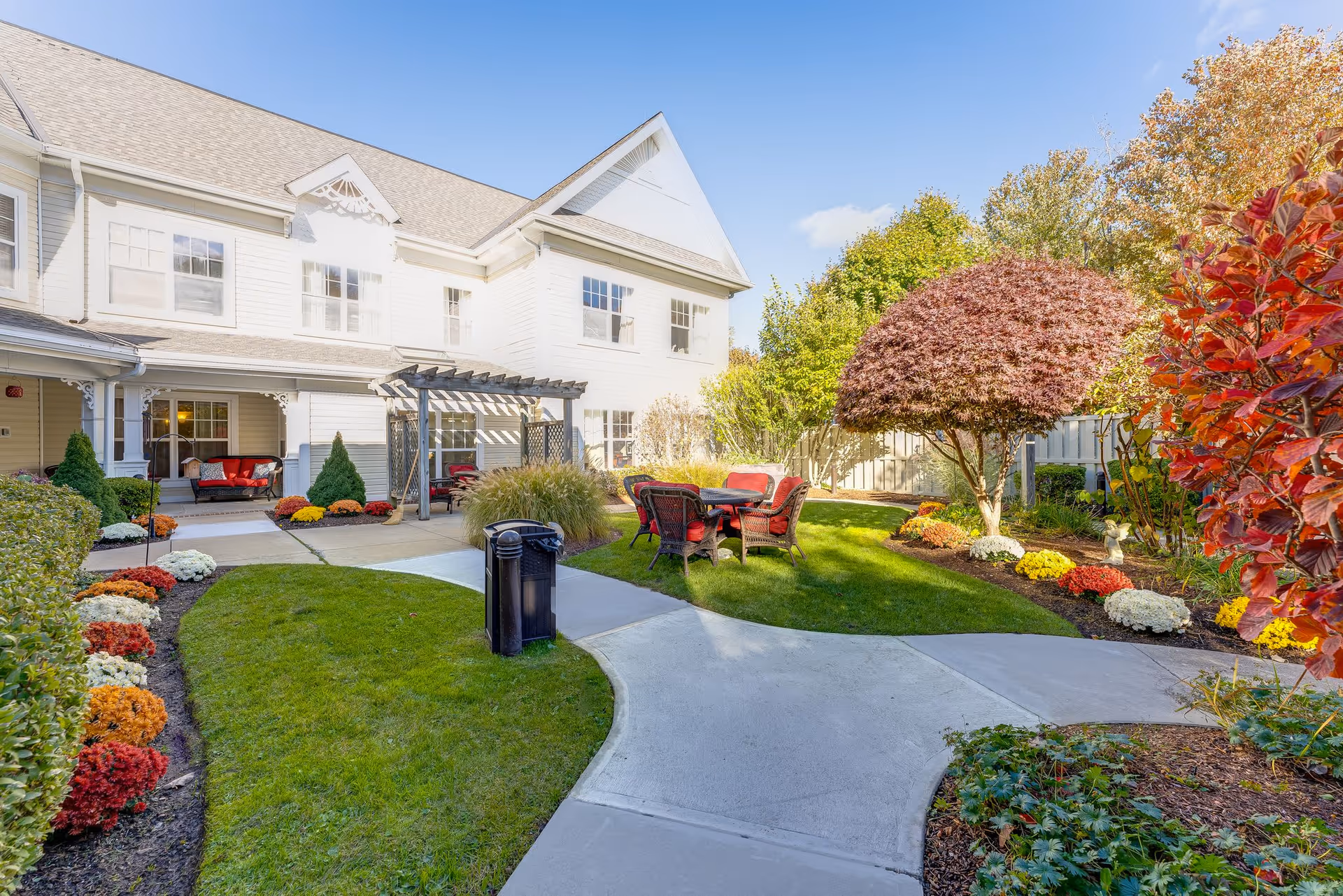Outdoor garden area at Sunrise of Wayland featuring a paved walkway, green lawn, colorful flower beds, a small tree with red leaves, and patio furniture with red cushions under a pergola and in the open air.