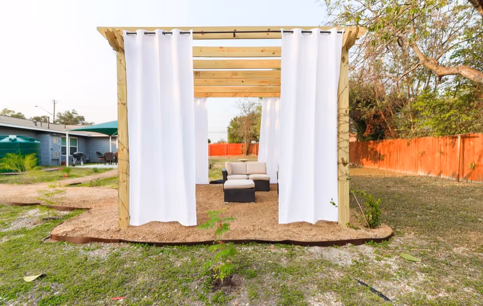 Outdoor wooden pergola structure with white curtains hanging on the sides, containing cushioned patio furniture including a sofa and ottoman, set on a gravel surface with grass and trees surrounding it.