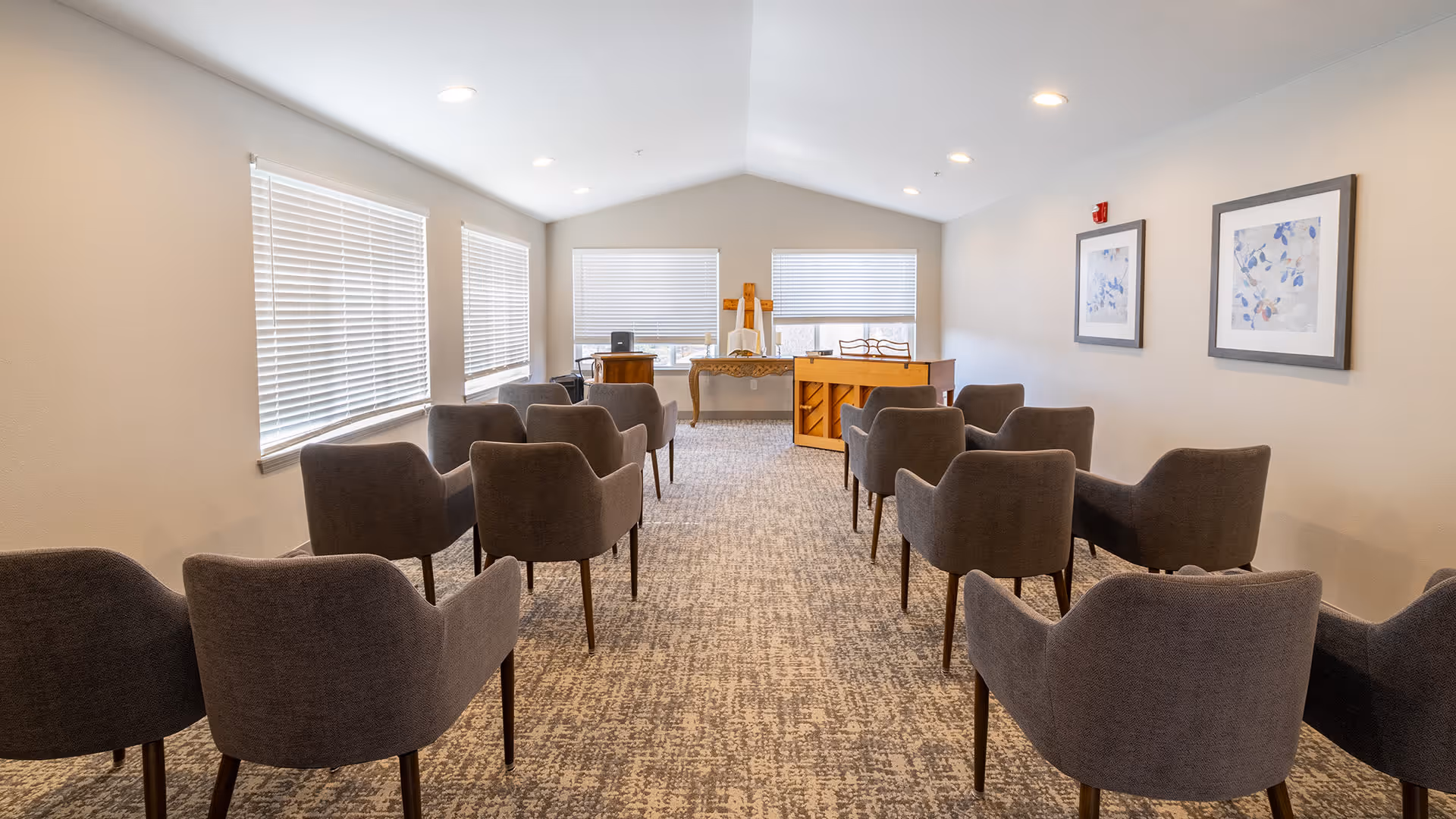 A small meeting or activity room with two rows of gray upholstered chairs facing a piano and a table at the front. The room has beige walls, three windows with white blinds, two framed abstract paintings on the right wall, and carpeted flooring.