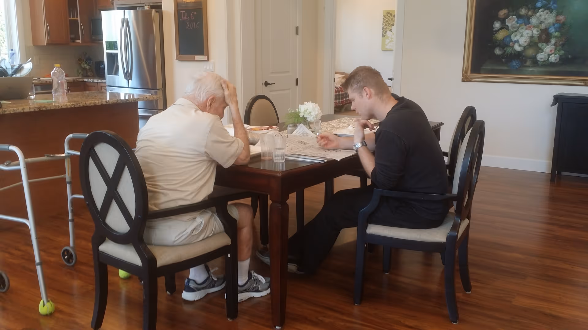 Two men sit at a dining table in a bright dining area with a walker nearby and a kitchen in the background.
