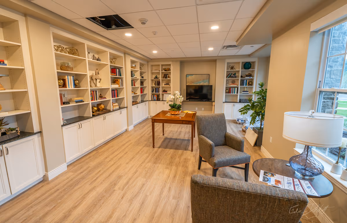 A bright and spacious living room area with light wood flooring, built-in white shelves filled with books and decorative items, a wooden table with a plant centerpiece, two gray upholstered chairs, a round side table with a lamp and magazines, and a flat-screen TV mounted on the far wall. Large windows allow natural light to fill the room.