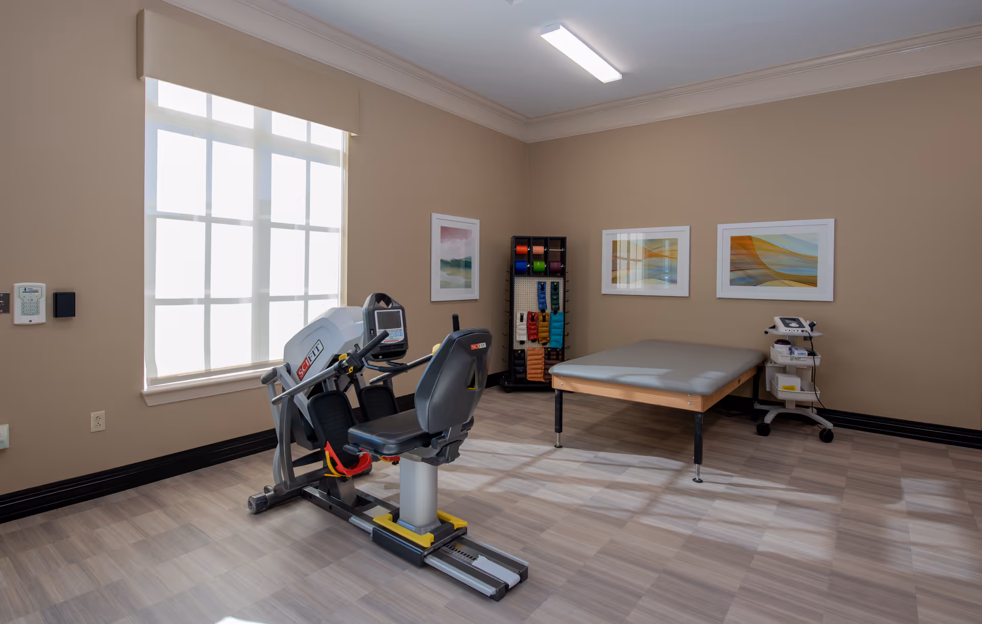 A well-lit therapy or exercise room with a large window covered by a white blind, a recumbent exercise bike, a padded therapy table, a rack with colorful resistance bands, and medical equipment on a small cart. The walls are beige with three abstract framed artworks.
