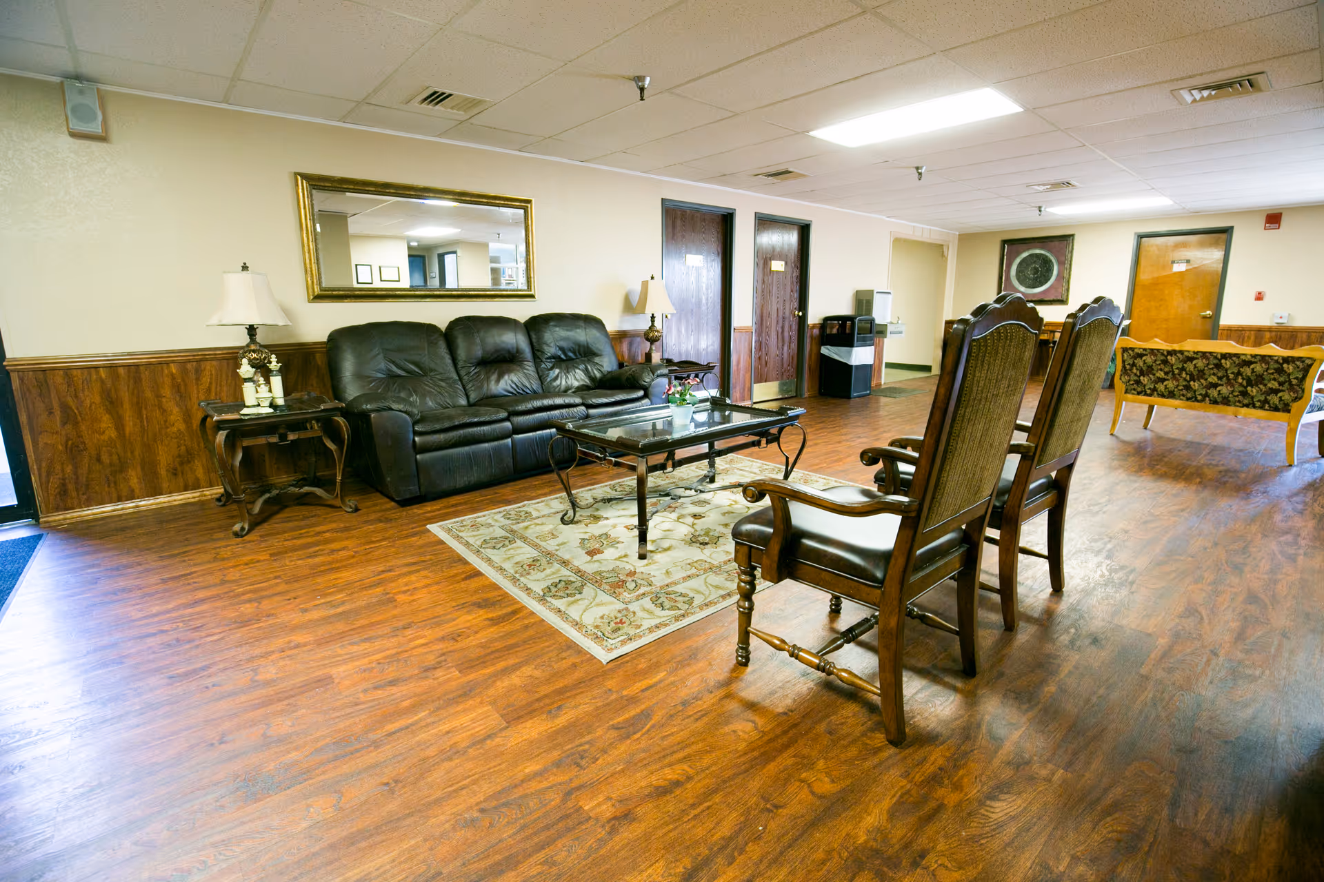Spacious senior living lounge with leather sofa, wooden chairs, coffee table, and warm wood flooring.