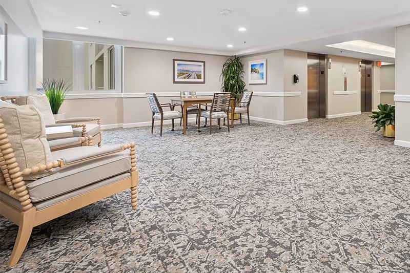 A spacious interior hallway area with patterned carpet, beige walls, and recessed lighting. There are two wooden armchairs with beige cushions on the left, a wooden table with four chairs in the center, and two framed pictures on the wall. To the right, there are two stainless steel elevators and a potted plant near the corner.