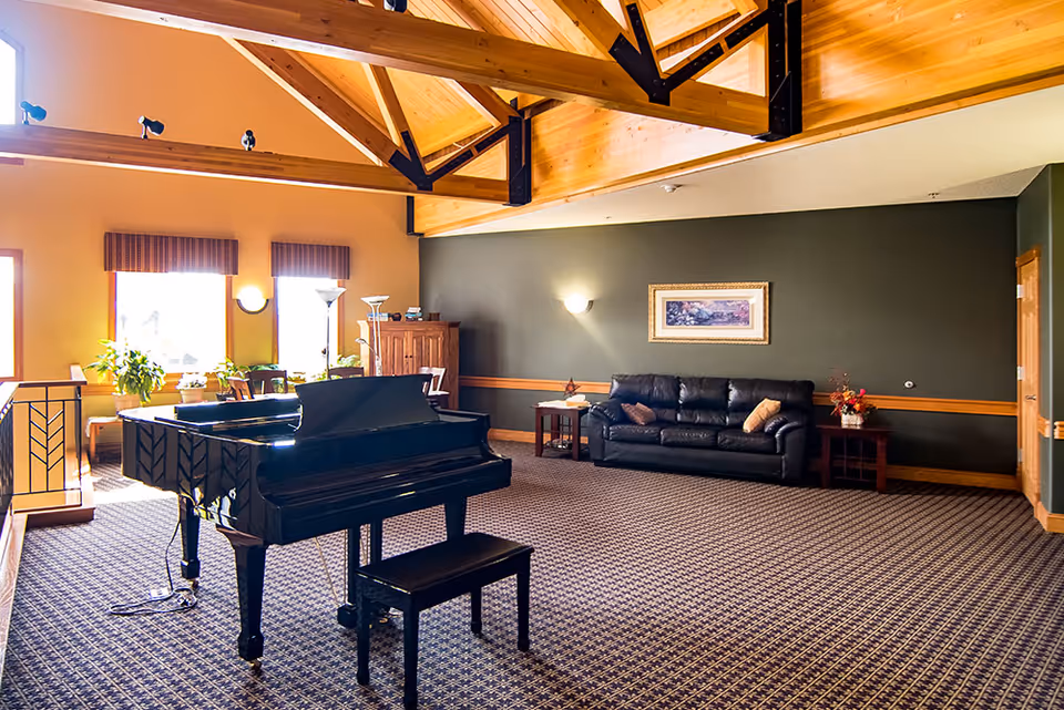 A spacious living room with a black grand piano and matching bench in the center. The room features a patterned carpet, a black leather couch with pillows against a dark green wall, two side tables with decorative items, and large windows letting in natural light. The ceiling has exposed wooden beams with black metal supports.