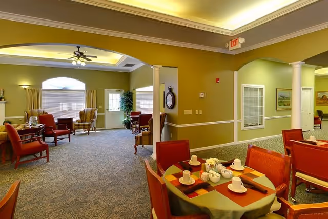 A spacious, well-lit common area in a senior living facility featuring multiple seating arrangements with armchairs and tables. The room has green walls, carpeted floors, and ceiling lights with a fan. A table in the foreground is set with cups, saucers, and a flower centerpiece. There are large windows with curtains and a door leading outside.