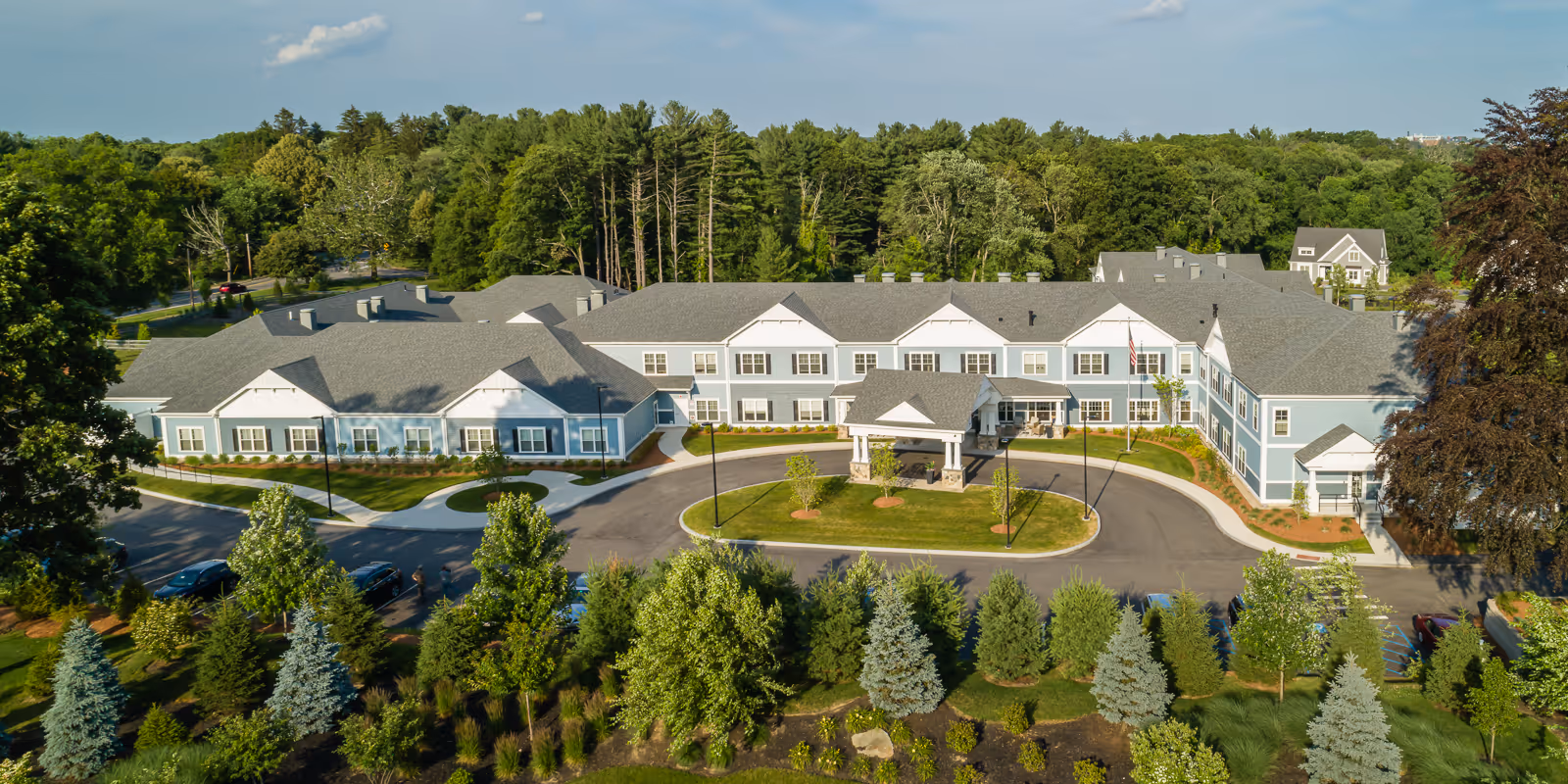 Aerial view of a large, two-story senior living facility building with light blue siding and white trim, surrounded by well-maintained landscaping including various trees and shrubs. The building has a covered entrance with a circular driveway and parking spaces in front. The background shows dense green trees under a partly cloudy sky.