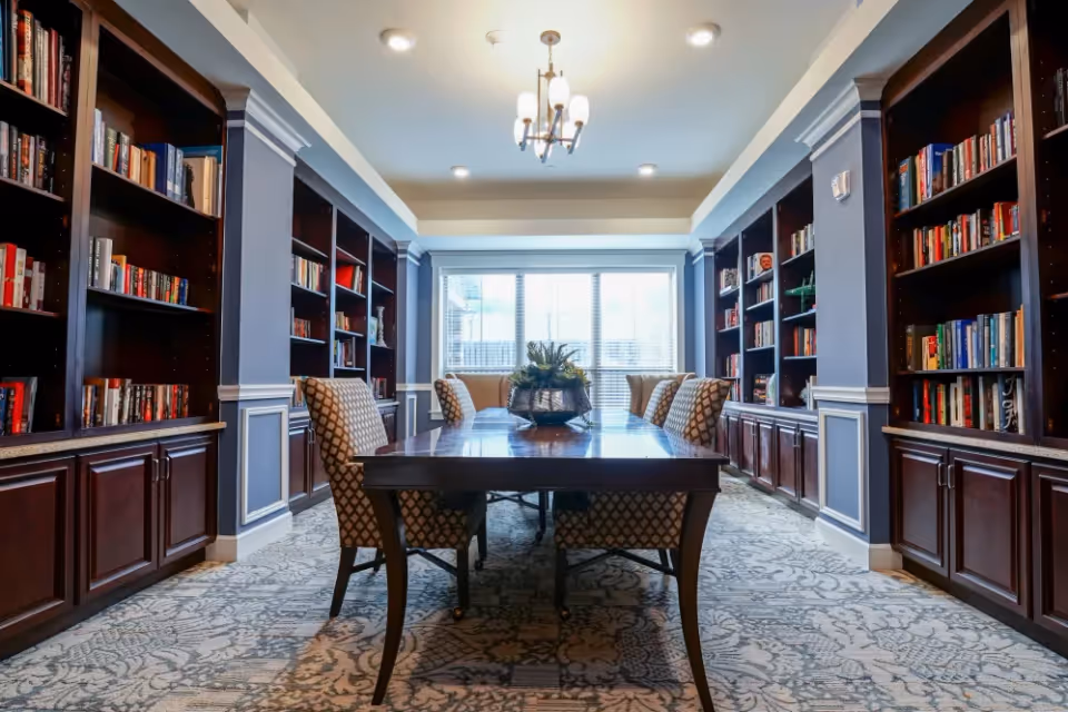 A well-lit library or reading room with dark wooden bookshelves lining both sides filled with books. In the center, there is a long wooden table with six patterned upholstered chairs around it. A decorative plant centerpiece is on the table. The room has a large window at the far end letting in natural light, and a chandelier hanging from the ceiling.