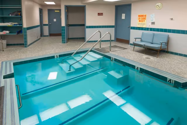 Indoor swimming pool with steps and handrails leading into the water. The pool area has tiled walls with teal accents, a seating bench, and several doors leading to other rooms. A sign on the wall reads 'WARNING NO LIFE GUARD ON DUTY'.