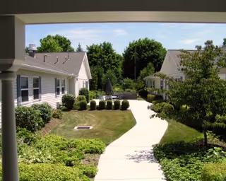 Sunlit courtyard with a paved walkway between single-story buildings, surrounded by shrubs, trees, and patio seating.