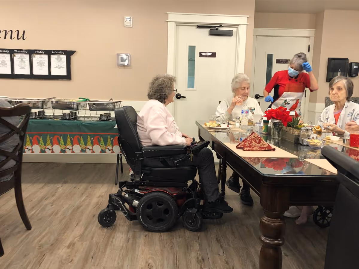 Several elderly women sitting around a dining table in a communal dining area. One woman is in a motorized wheelchair, and a staff member wearing a mask and gloves is standing nearby. The table has placemats, a red poinsettia centerpiece, and various food items. In the background, there is a buffet table with covered trays and a menu on the wall.