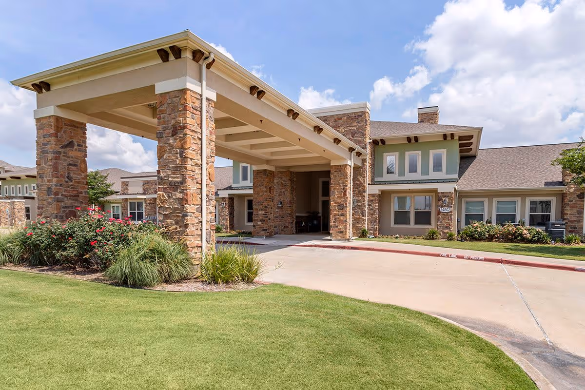 Exterior view of Caydance Assisted Living & Memory Care facility showing a covered entrance supported by stone pillars, manicured lawn, and a clear blue sky with some clouds.
