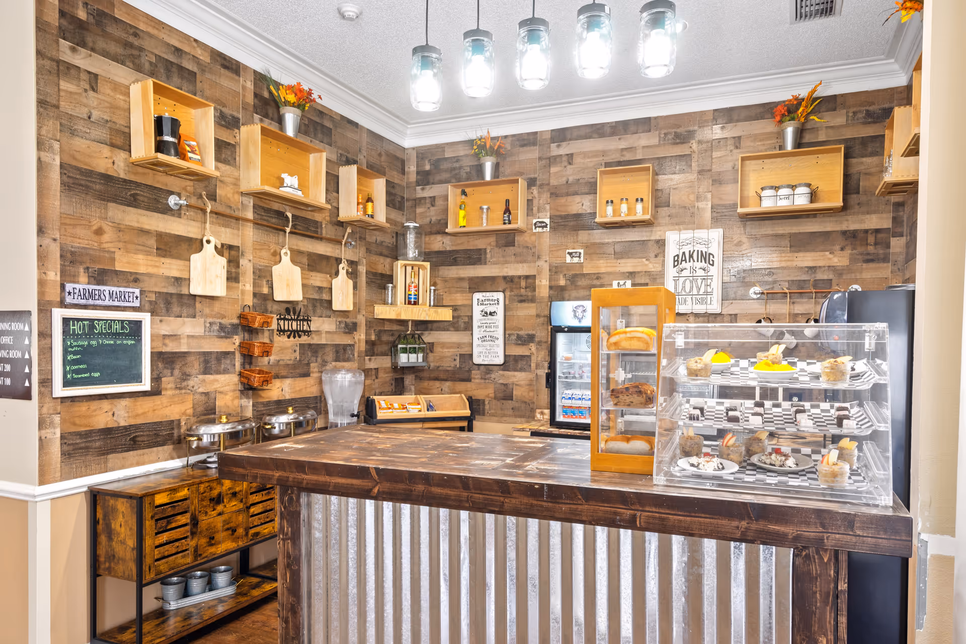 A rustic-style kitchen area with wooden walls and shelves holding various jars, bottles, and decorative items. A wooden counter with a corrugated metal front is in the foreground, displaying baked goods in glass cases. The wall features signs including a chalkboard with hot specials and decorative plaques. Overhead, there are five hanging lights with glass covers.