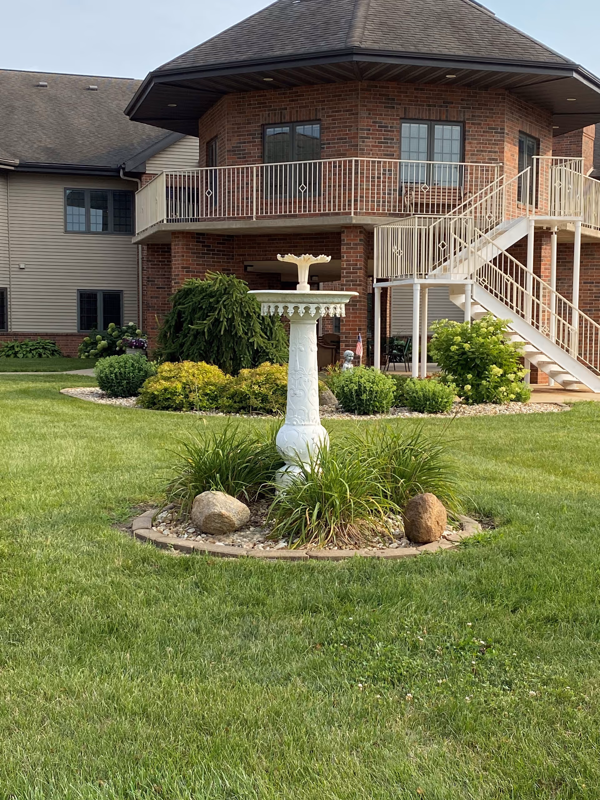 A landscaped outdoor area with a white decorative birdbath surrounded by grass, plants, and rocks in the foreground. In the background, there is a two-story brick and siding building with a balcony and an exterior staircase.