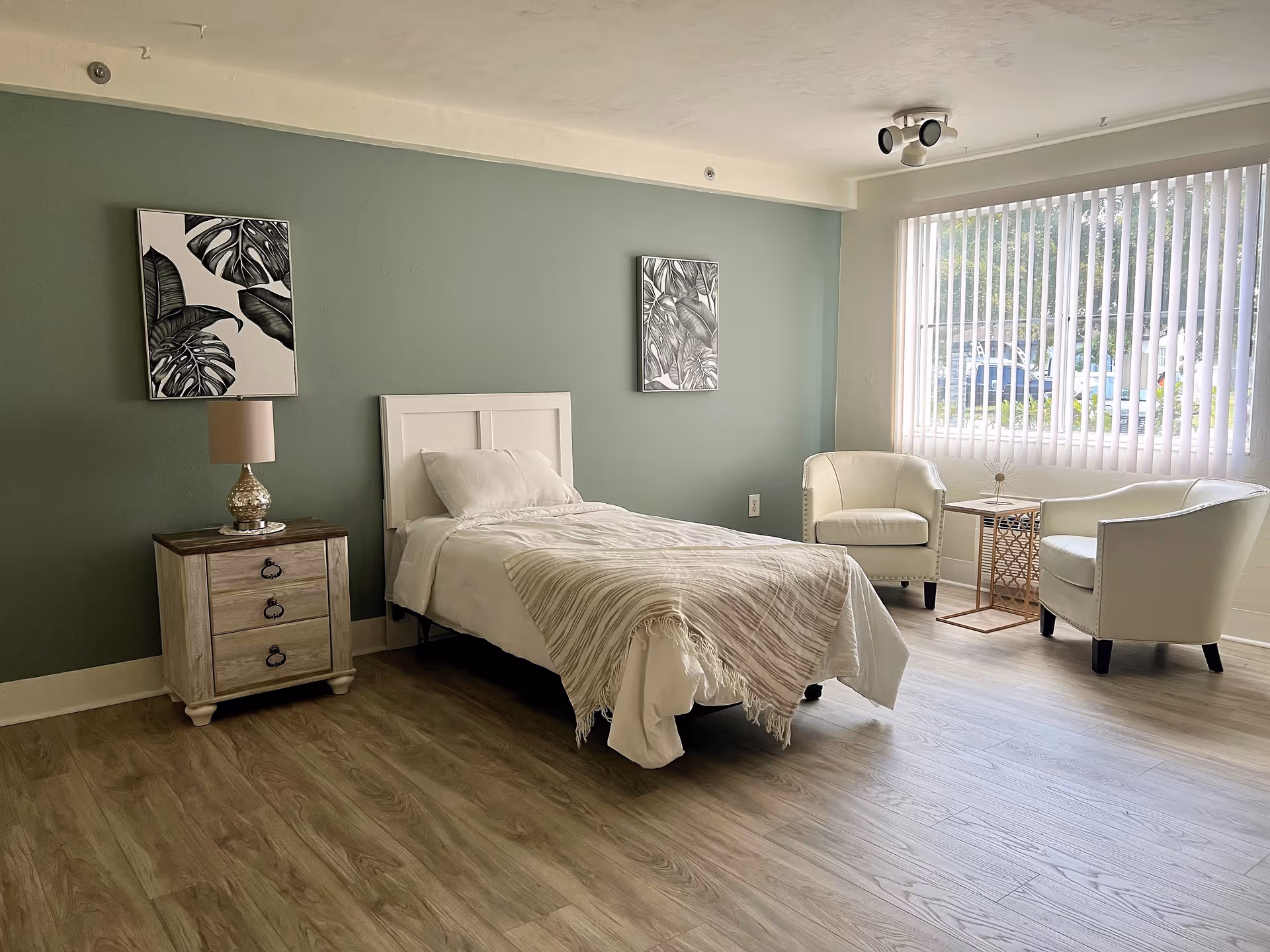 A neatly arranged bedroom with a single bed covered in white bedding and a beige throw blanket. Next to the bed is a wooden nightstand with a decorative lamp. Two framed botanical prints hang on a green accent wall behind the bed. On the right side of the room, there are two white armchairs with a small decorative table between them, positioned near a large window with vertical blinds letting in natural light. The floor is covered with light wood laminate.