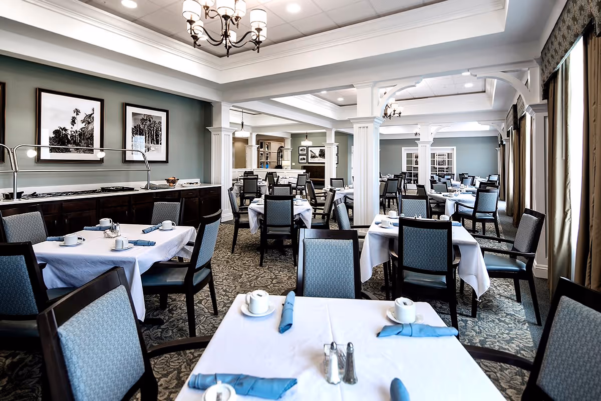 A spacious dining room with multiple tables covered in white tablecloths, each set with blue napkins, cups, and saucers. The room features patterned carpet, light blue walls with framed black and white photographs, white columns, and chandeliers hanging from the ceiling. Large windows with curtains allow natural light to enter the room.