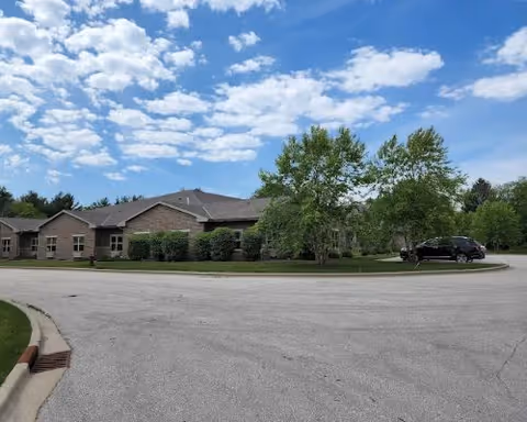 Single-story brick senior living building with a curved driveway, landscaping, and a parked vehicle under a partly cloudy sky.