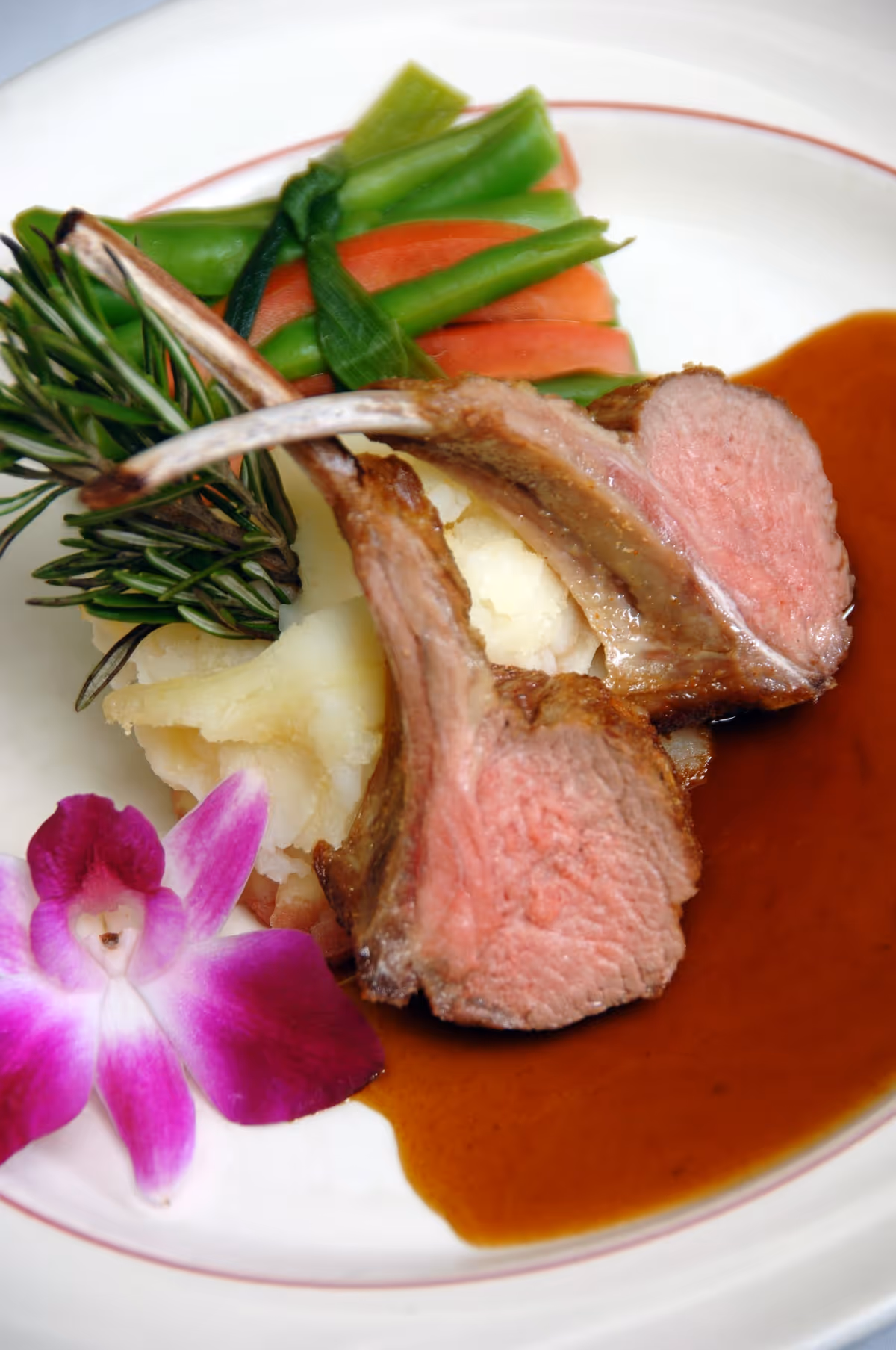 Plated lamb chops with mashed potatoes, green beans, brown sauce and a purple orchid garnish.