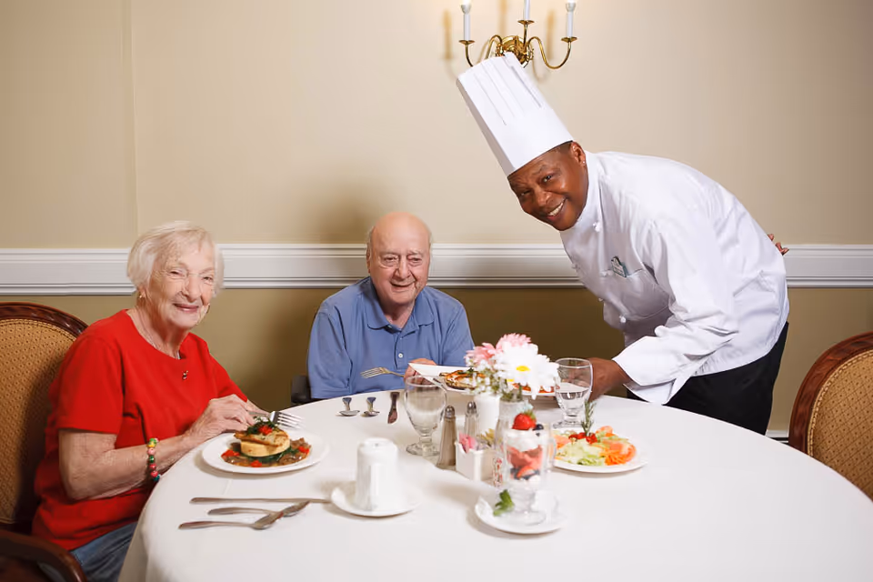 Two elderly residents sit at a dining table while a smiling chef serves their meal in a dining room.