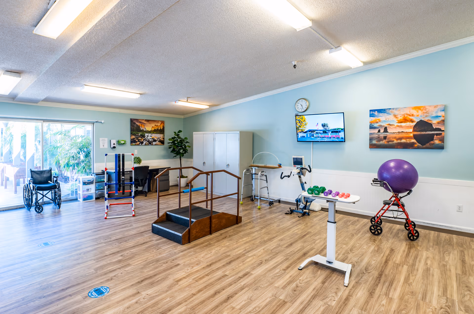 A spacious rehabilitation room with wooden flooring and light blue walls, featuring exercise equipment including a wheelchair, walker, stationary bike, small hand weights on a white table, a purple exercise ball on a red walker, and a set of stairs with handrails for physical therapy. The room has large windows letting in natural light, a wall-mounted TV, a clock, and colorful landscape paintings on the walls.