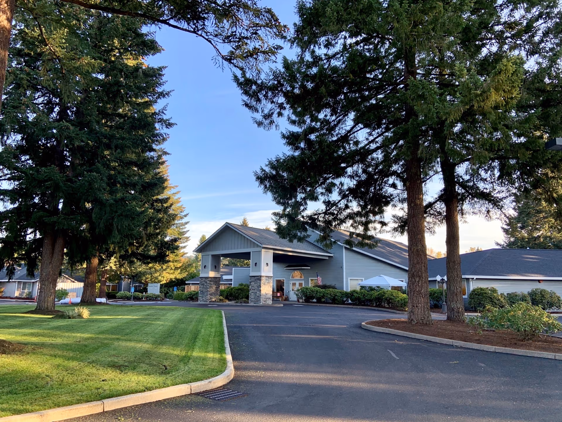 Exterior view of Magnolia Gardens Senior Living facility with a driveway leading to the entrance. The building has a covered entryway supported by stone pillars. Tall evergreen trees and well-maintained green lawns surround the area under a clear blue sky.