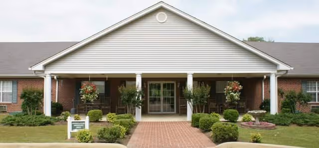 Front exterior view of a single-story senior living facility building with a covered entrance supported by white columns, brick walls, and a gabled roof. There are shrubs, hanging flower baskets, and a brick walkway leading to the entrance.