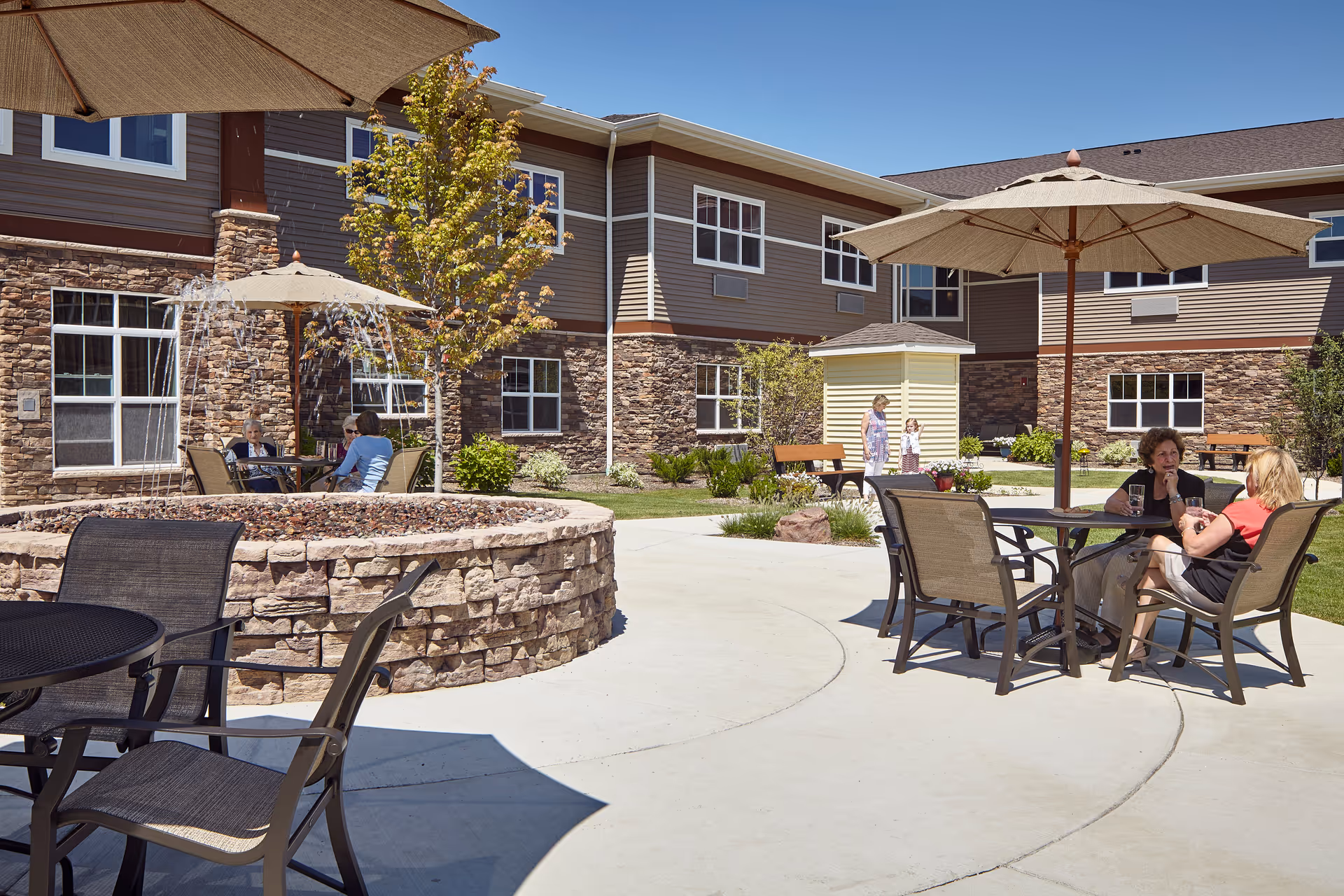 Outdoor courtyard area at Terra Vista of Oakbrook Terrace with stone walls, patio tables with umbrellas, and people sitting and conversing. The building exterior features stone and siding with multiple windows. There is a small fountain and some greenery around the courtyard.