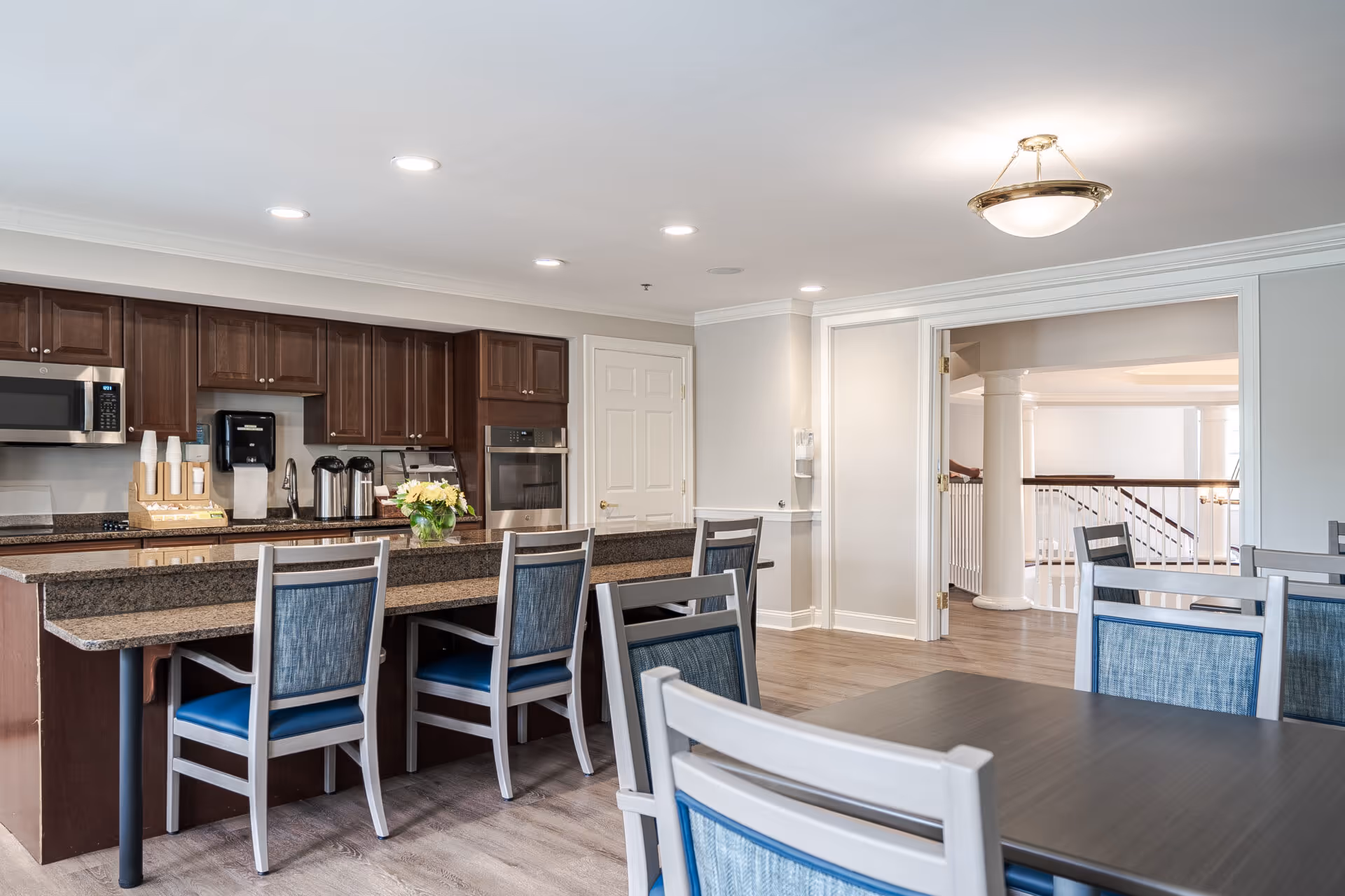 A modern kitchen and dining area in a senior living facility featuring dark wood cabinets, granite countertops, stainless steel microwave and oven, coffee station with cups, and several chairs around a kitchen island and dining tables. The room has recessed lighting and a hanging light fixture, with an open doorway leading to a hallway with white railings and columns.